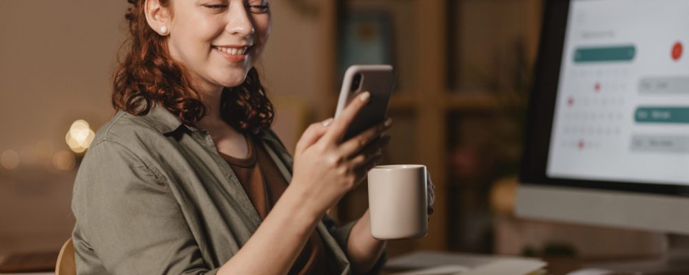 mulher-em-casa-usando-smartphone-na-frente-computador-enquanto-toma-cafe